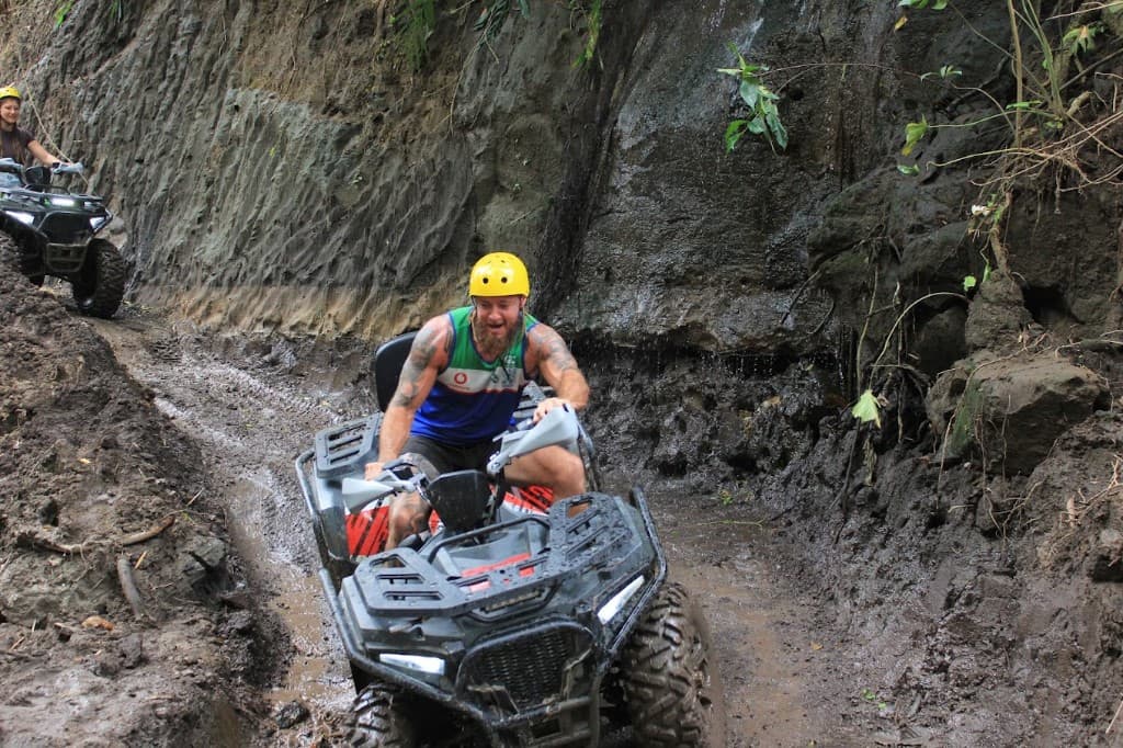 A smiling rider in a yellow helmet driving a grey quad along a muddy jungle track beside a dripping rock wall