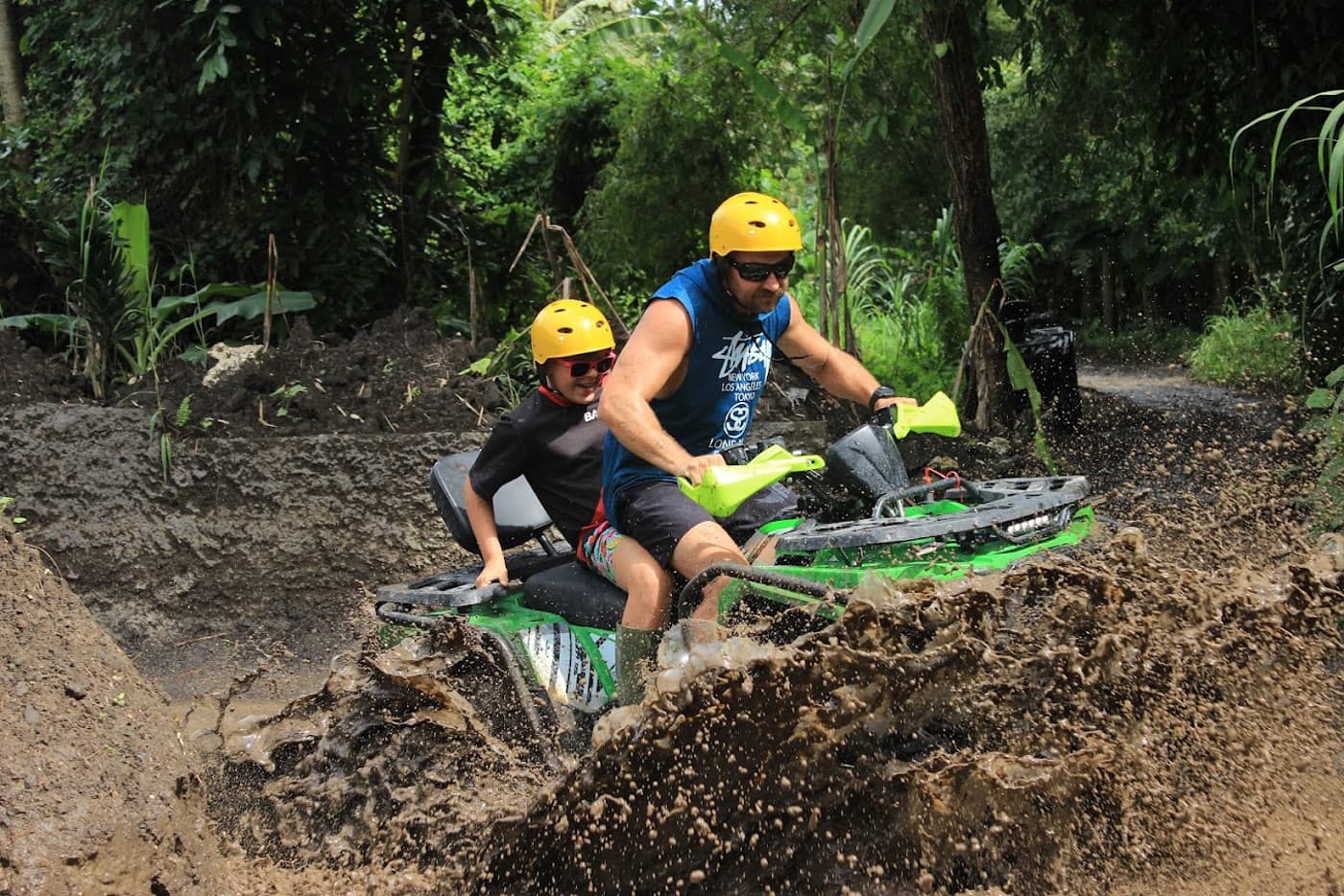 Two riders in yellow helmets sharing one green quad through jungle mud