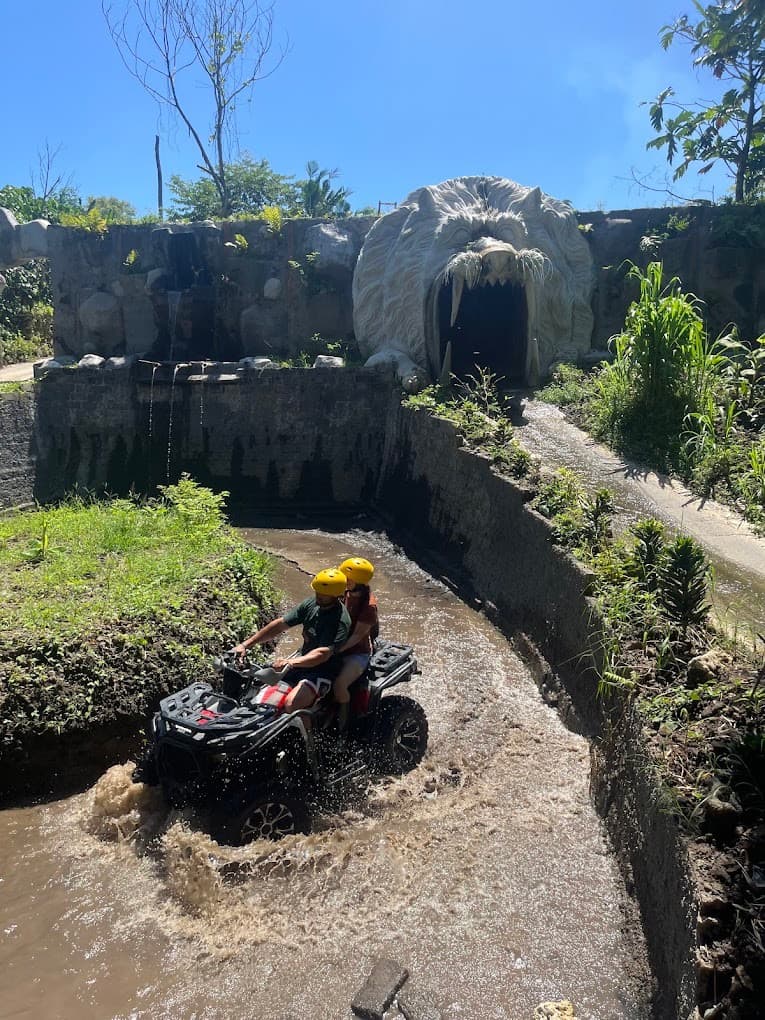 Two riders on one quad in the river channel below a tunnel and waterfall