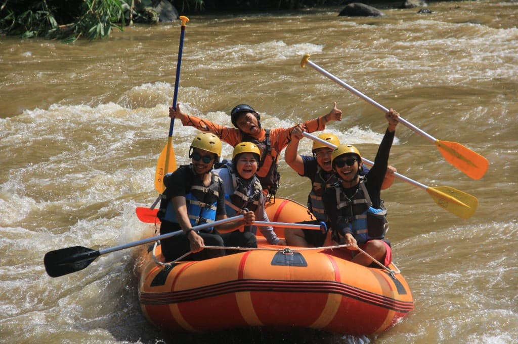 First-time rafters in yellow helmets and life jackets cheering with paddles raised in an orange raft on the Ayung