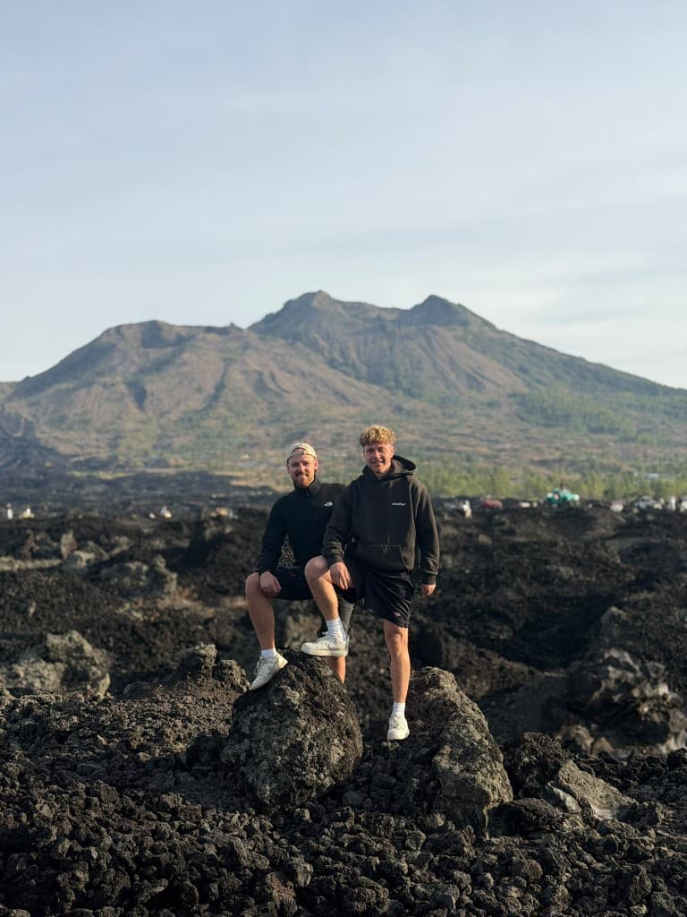 Two guests standing on black lava rocks with the cone of Mount Batur behind them