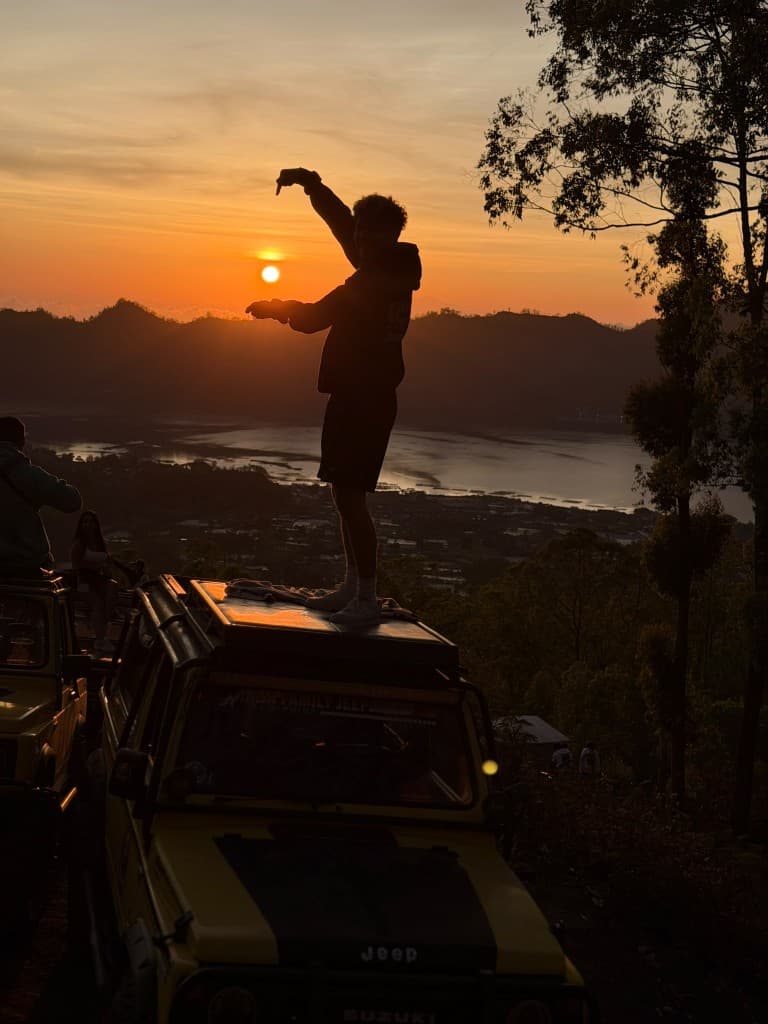A guest in silhouette on a jeep roof framing the Batur sunrise with both hands