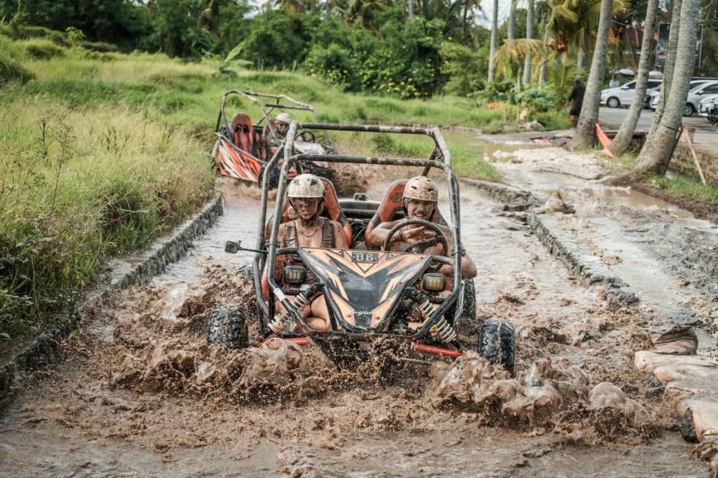 A driver and passenger in helmets splashing along a muddy channel beside rice fields in an open two-seat buggy, another buggy behind