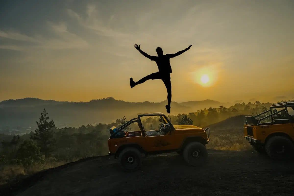 A person jumping in front of an open jeep at sunrise above a mountain caldera, with a second jeep beside it