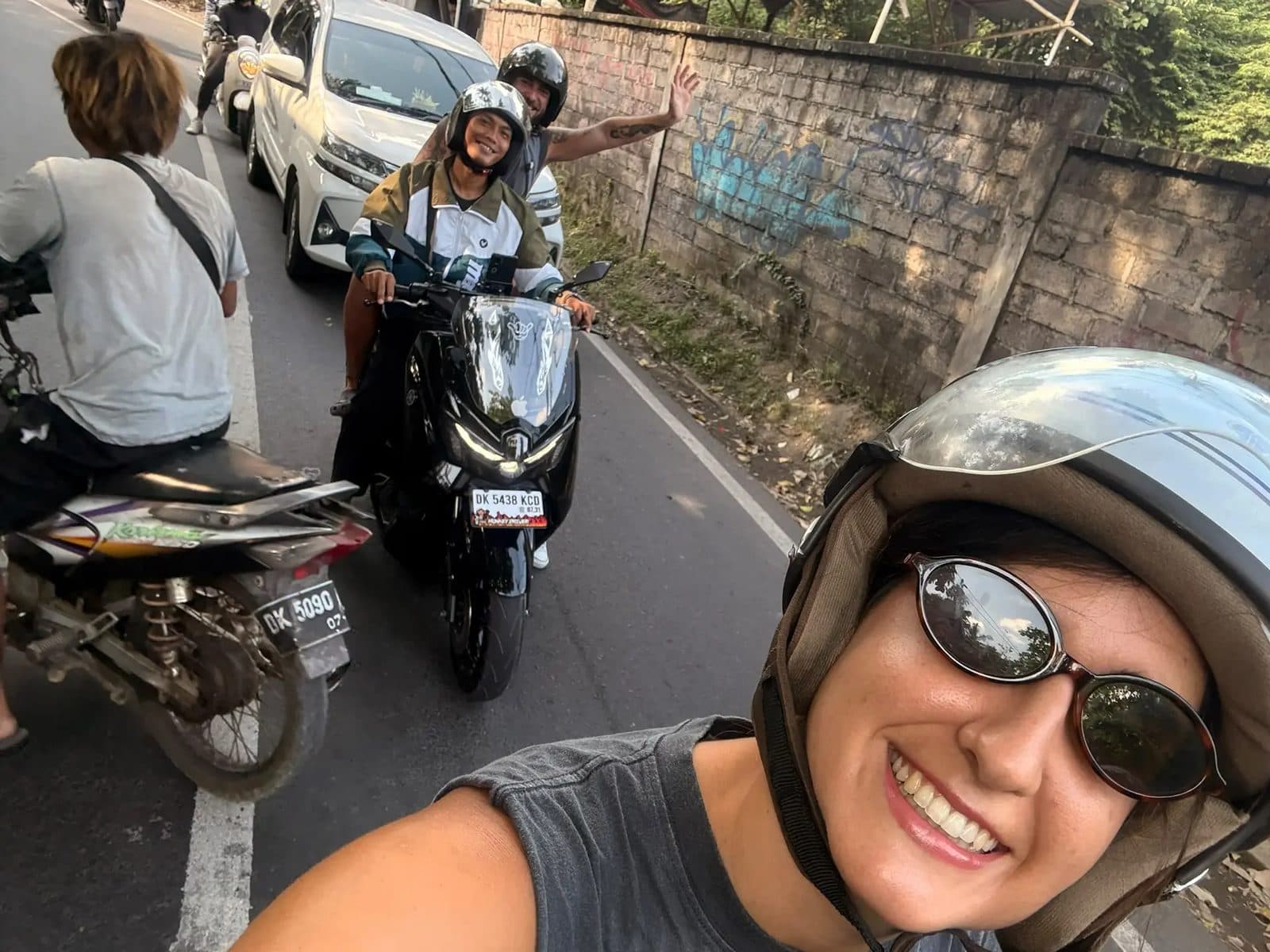 Riders on automatic scooters on a village road, helmets on, one person waving from the scooter behind